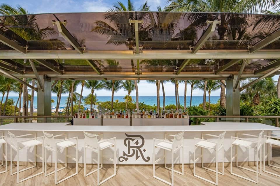Beachfront bar with white chairs, periscope ceiling, palm trees, and ocean view in background