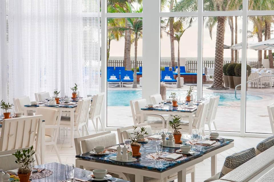 Upscale dining room with white chairs, ocean-view windows, and resort pool visible outside