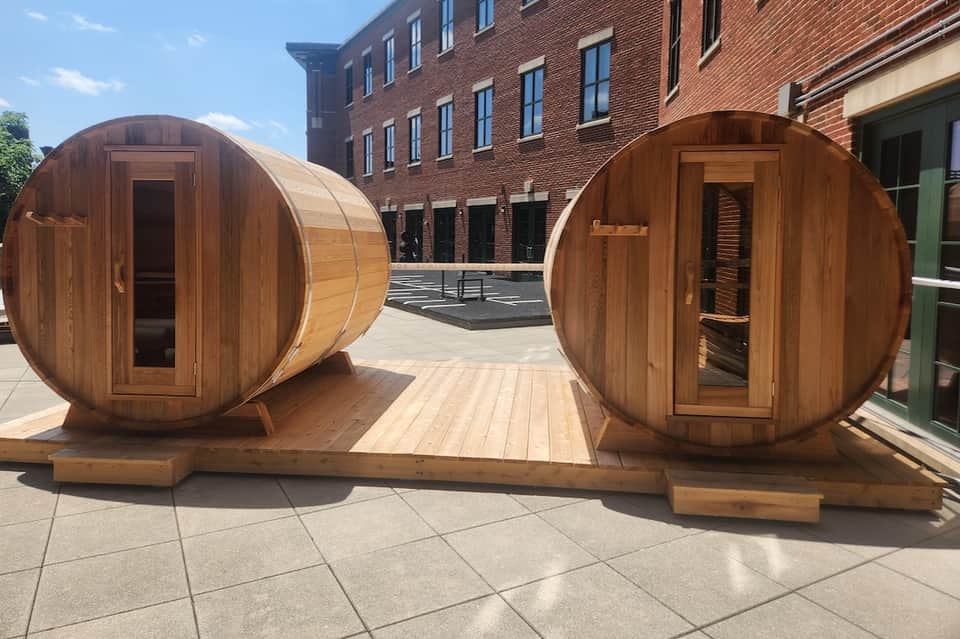 Two wooden barrel saunas on outdoor patio with brick buildings