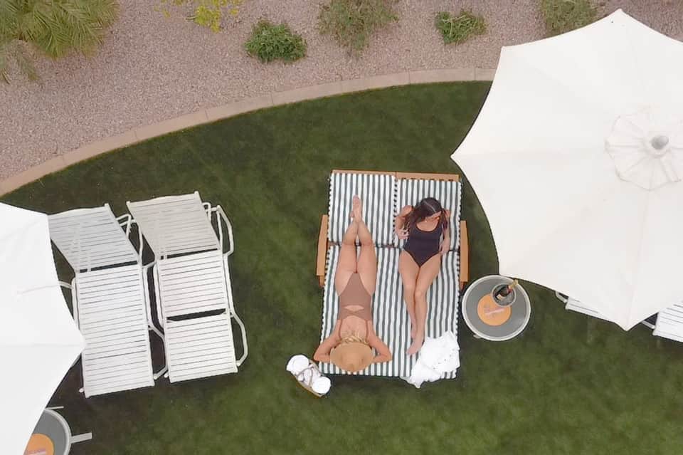 Aerial view of two women on lounge chairs on grass under white umbrellas next to resort landscaping