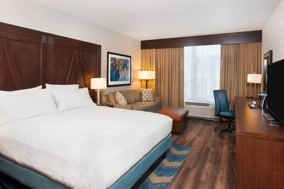 Hotel bedroom with queen bed, dark wood headboard, seating area, and city window views