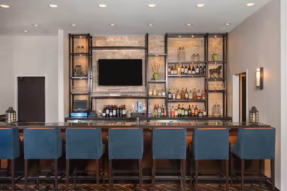 Modern hotel bar with illuminated shelving displaying bottles, TV screen, and blue upholstered seating