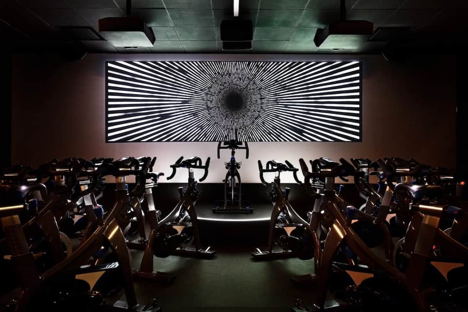 Indoor spin studio with rows of exercise bikes facing large illuminated geometric wall display screen