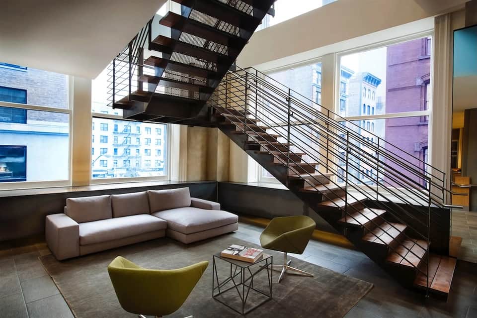 Industrial loft-style lobby with floating staircase, metal railings, large windows overlooking city skyline, and modern seating