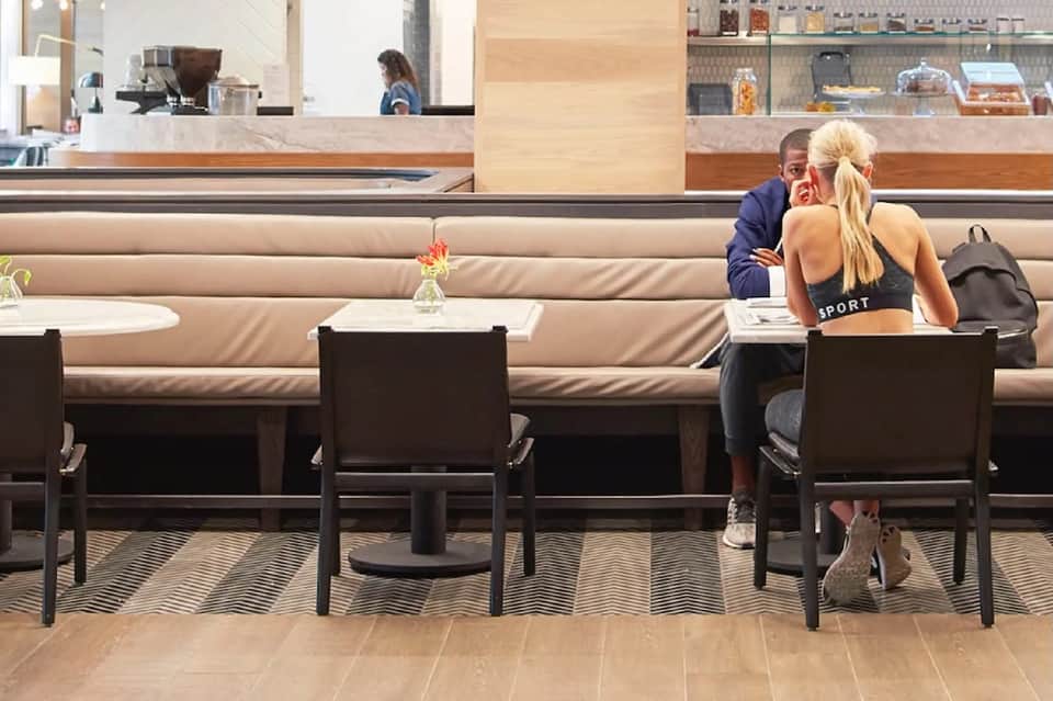 Modern hotel café with long beige banquette seating, black chairs, counter service, and minimalist design