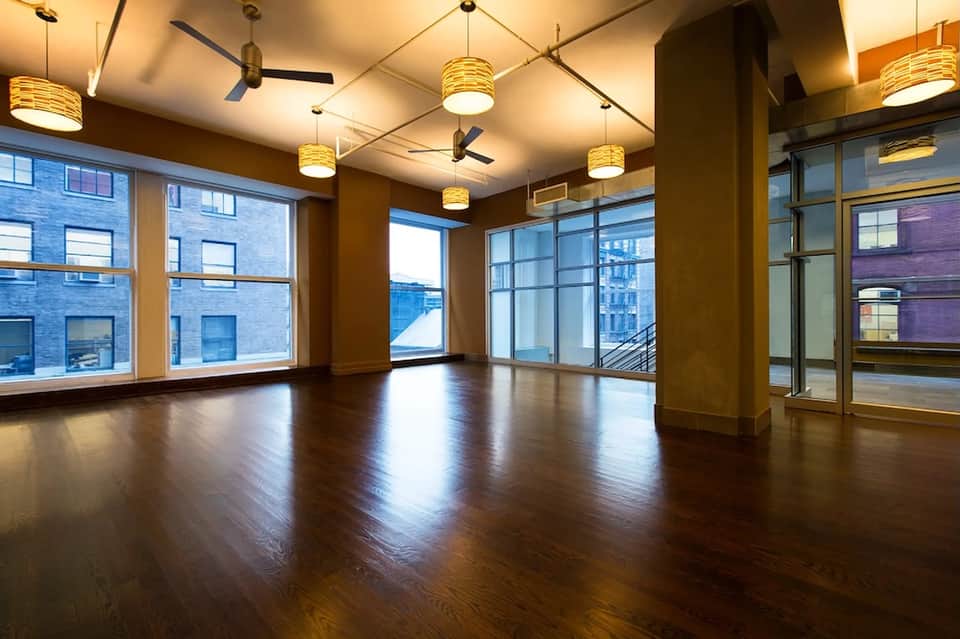 Empty modern loft space with dark wood floors, columns, pendant lights, and large windows