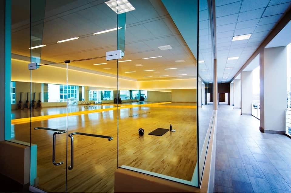 Modern fitness studio with mirrored walls, wooden floor, glass doors, yellow lighting, and city views