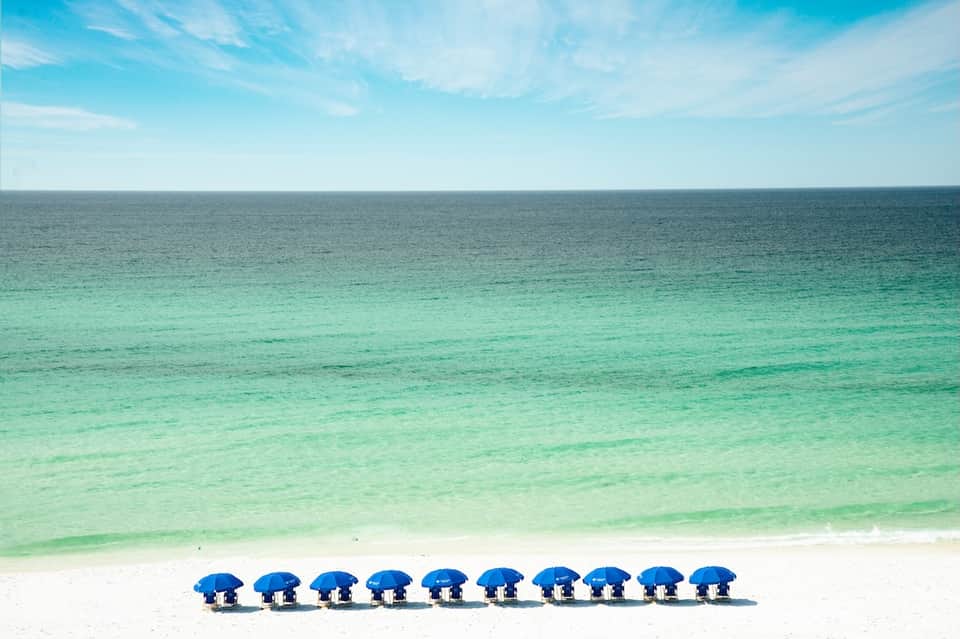 Beach scene with ten blue umbrellas lined up on white sand facing turquoise ocean