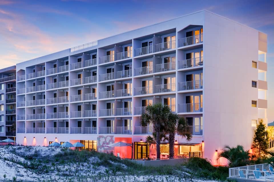 Modern beachfront hotel with illuminated facade at dusk, palm tree, and sandy dunes