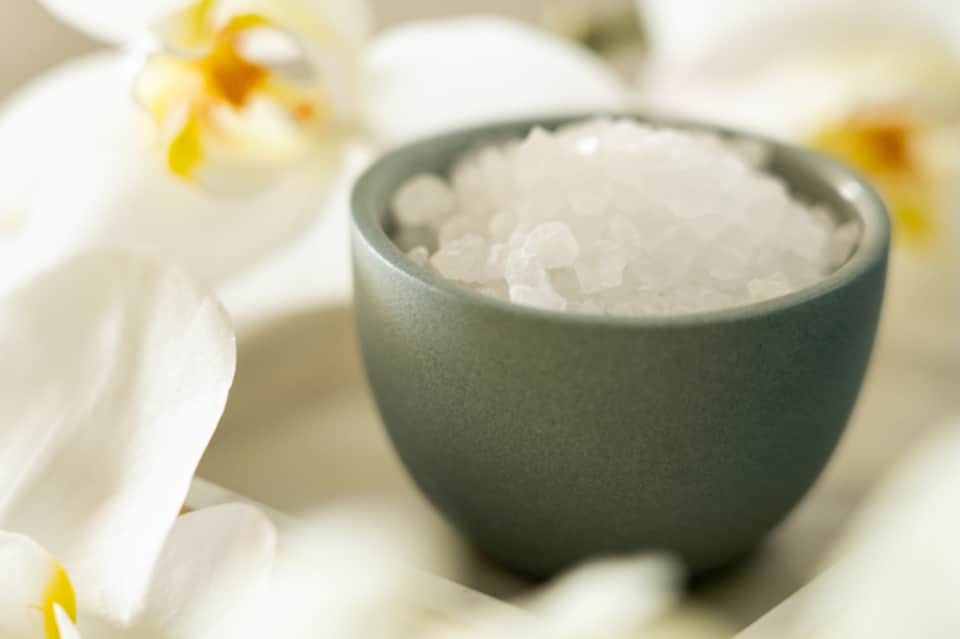 Green ceramic bowl filled with sea salt surrounded by white spa flowers on neutral background