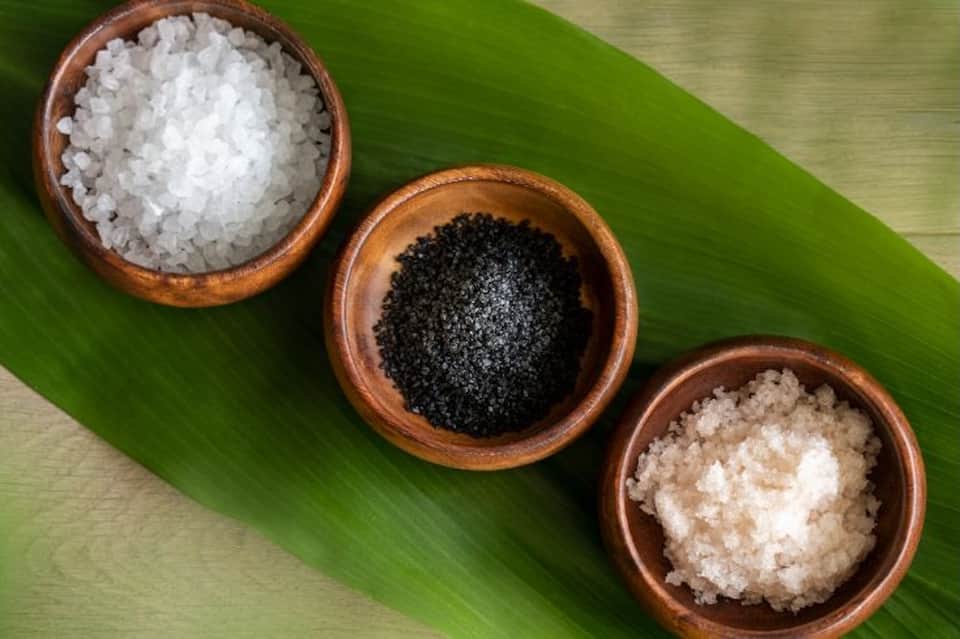 Three wooden bowls containing white salt, black salt, and pink salt on green leaf backdrop