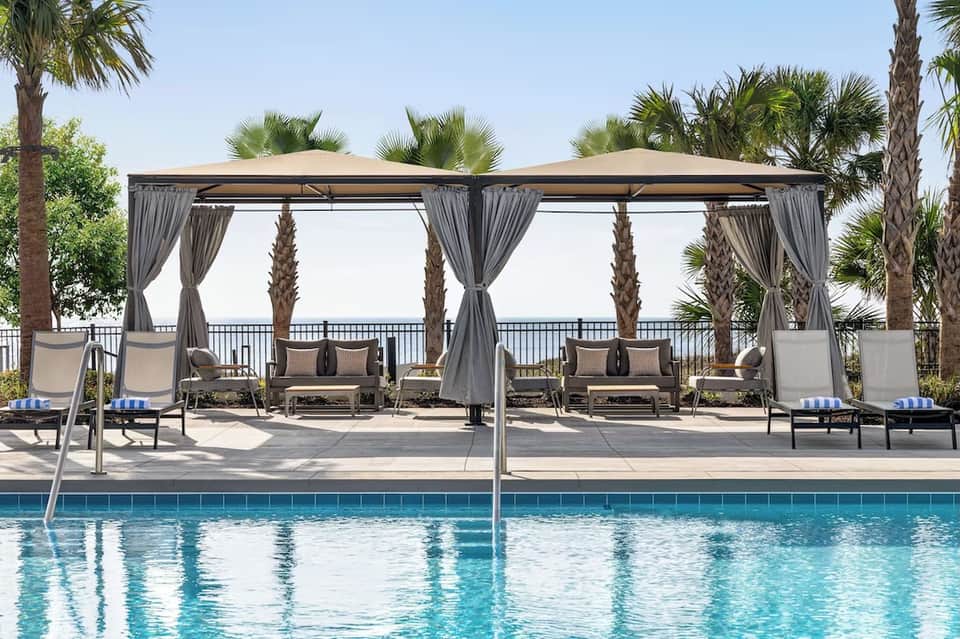 Resort pool with dual gray pergolas, lounge seating, palm trees, and ocean waterfront railing