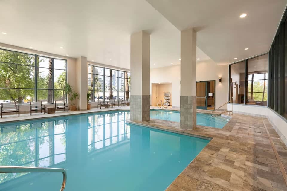 Indoor heated swimming pool with turquoise water, columns, large windows, and lounge seating area