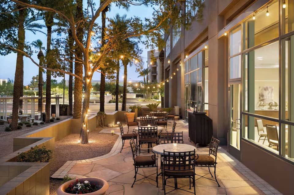 Evening courtyard patio with palm trees, bistro seating, warm lighting, fire pit, and modern building facade