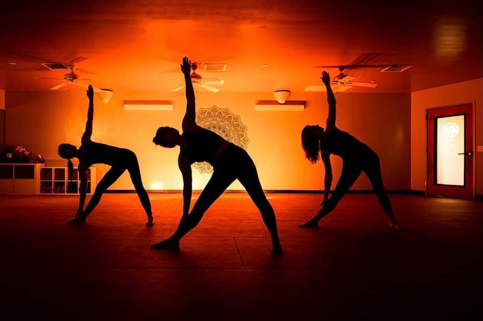 Fitness studio with orange lighting, three people performing yoga stretches on wooden floor