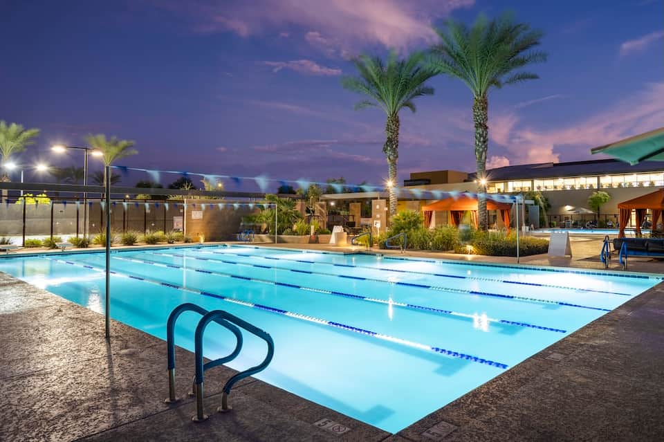 Lap pool at dusk with lane dividers, palm trees, modern facility buildings, and evening ambient lighting