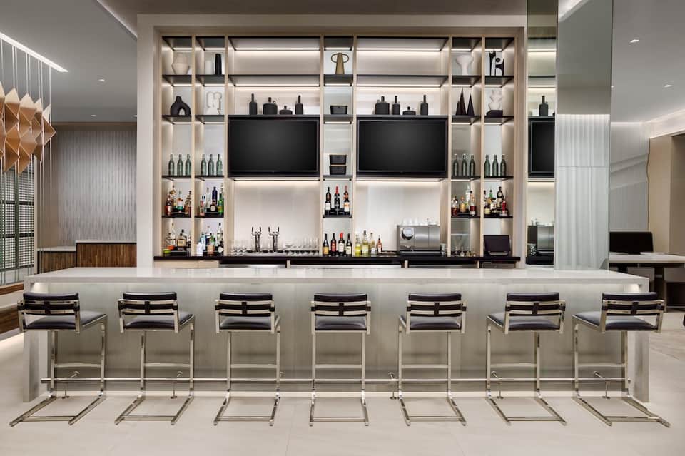 Contemporary hotel bar with white shelving, bottles of wine and spirits, flat-screen TVs, and black bar stools