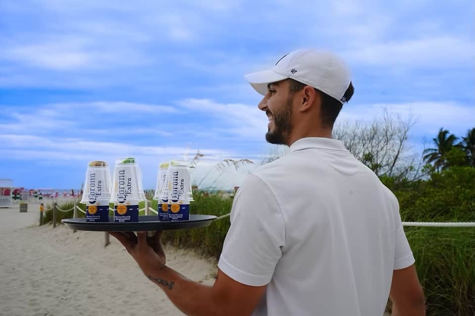 Server carrying Corona beer on beach with ocean and dunes in background at resort