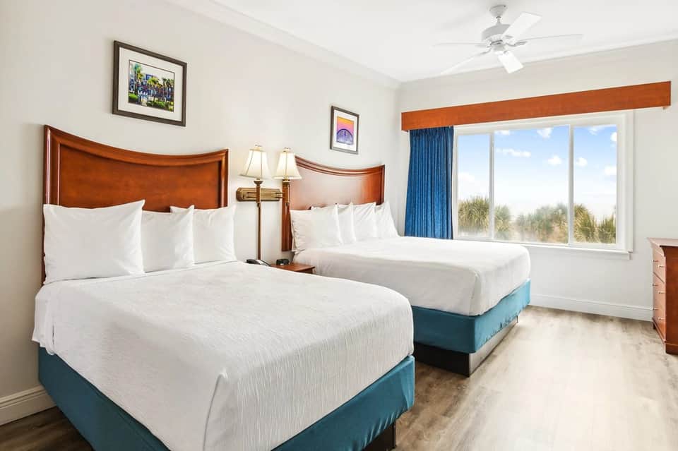 Hotel bedroom with two queen beds, wooden headboards, ocean view windows, and blue curtains