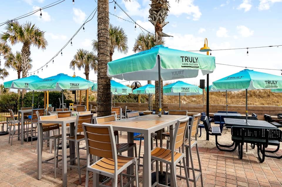 Beachside dining patio with turquoise umbrellas, wooden tables, chairs, palm trees, and string lights
