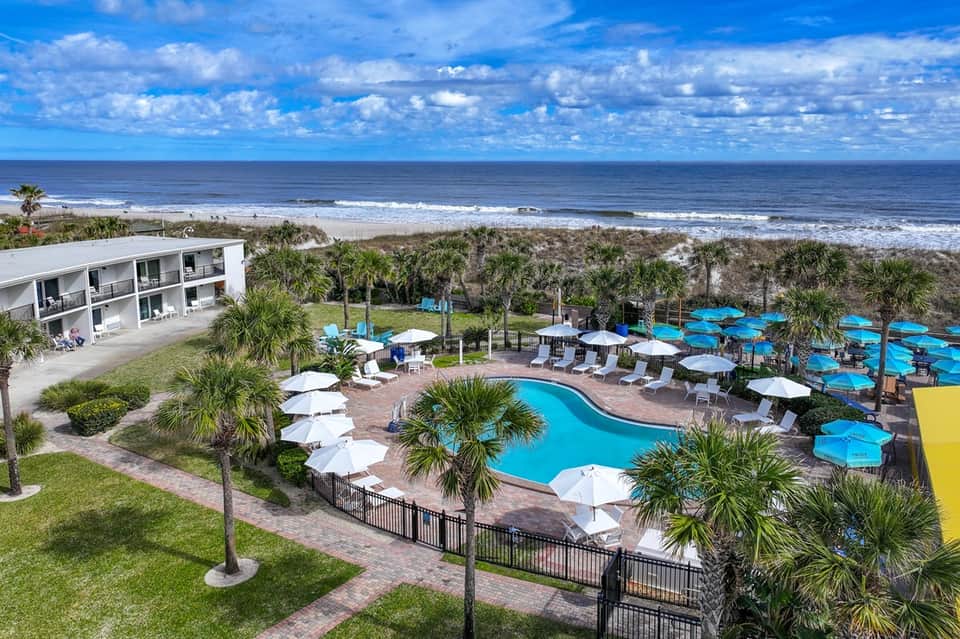 Beachfront resort pool with curved layout, white umbrellas, palm trees, and ocean view