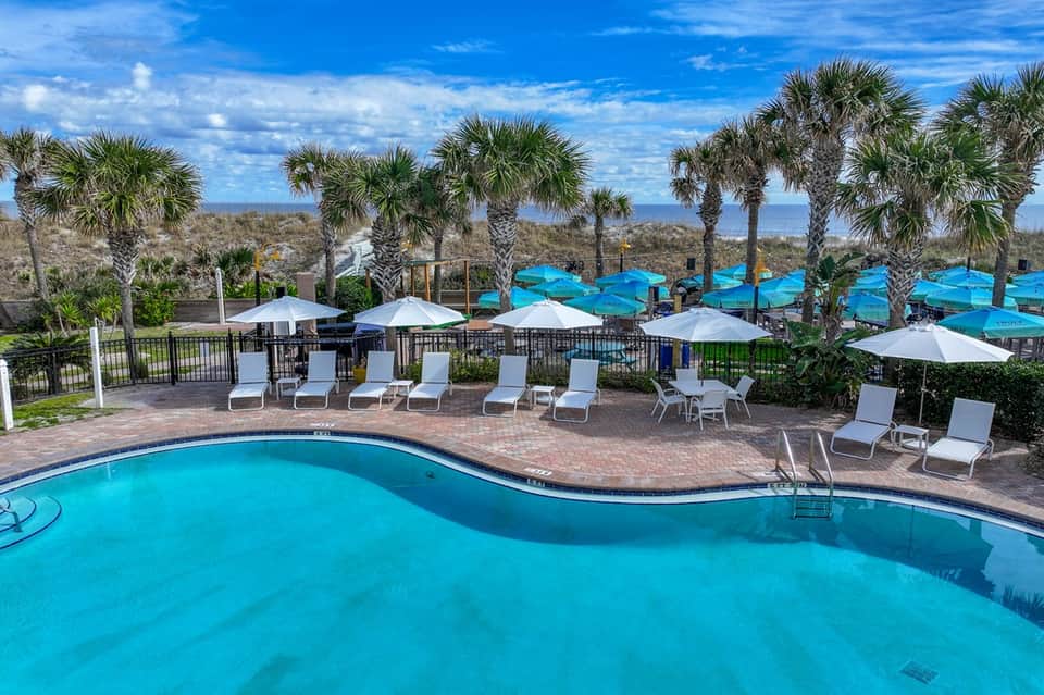 Scenic resort pool with curved edge, palm trees, blue umbrellas, and ocean view in background
