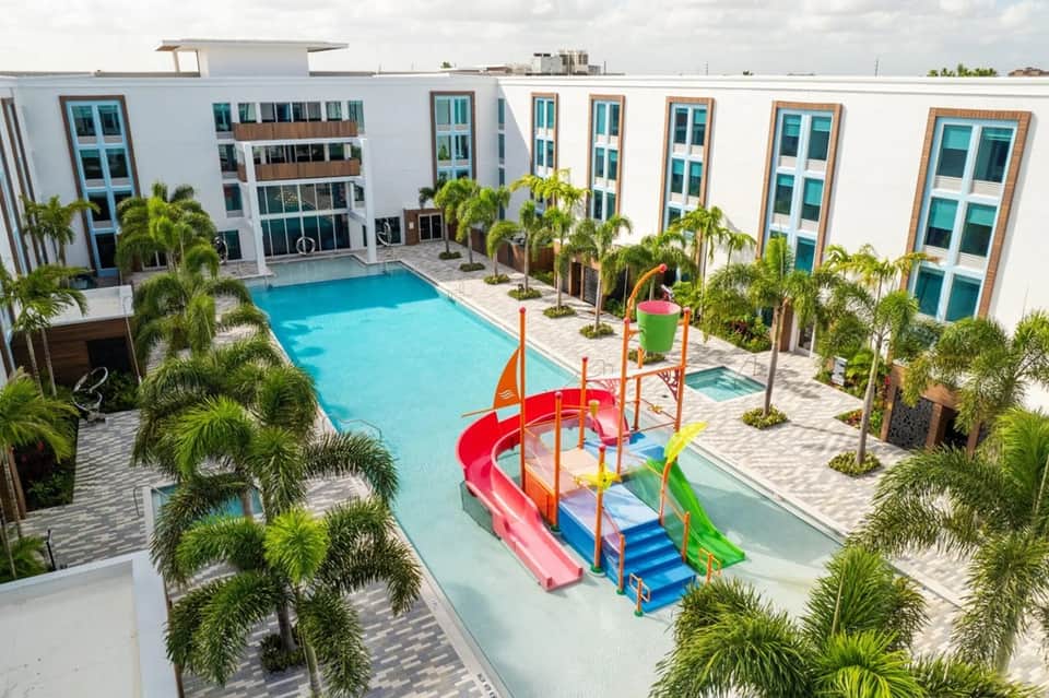 Resort courtyard with bright blue swimming pool, colorful waterslide, palm trees, white hotel buildings, and paved grounds