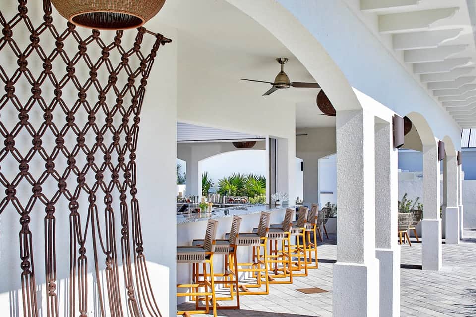 Modern resort covered patio with macramé wall hanging, bar seating, and ocean view beyond columns