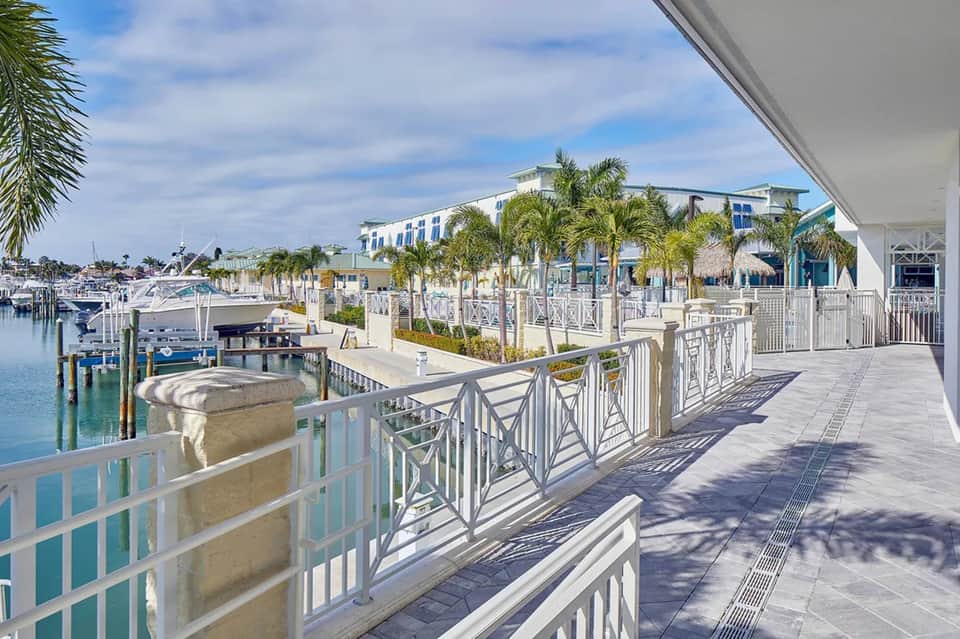 Waterfront boardwalk with white railings, marina boats, palm trees, and modern coastal buildings