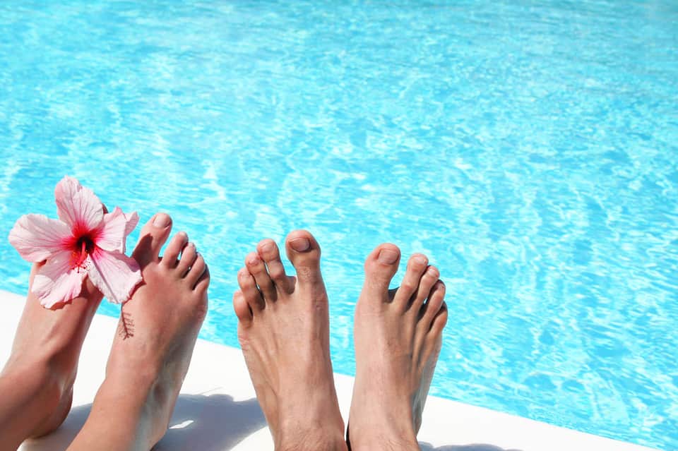 Four people's feet dangling poolside with pink hibiscus flower and turquoise water below