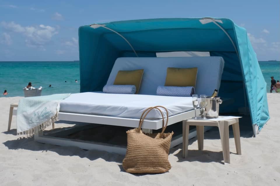 Private beach cabana bed with turquoise canopy overlooking ocean with white cushioned seating