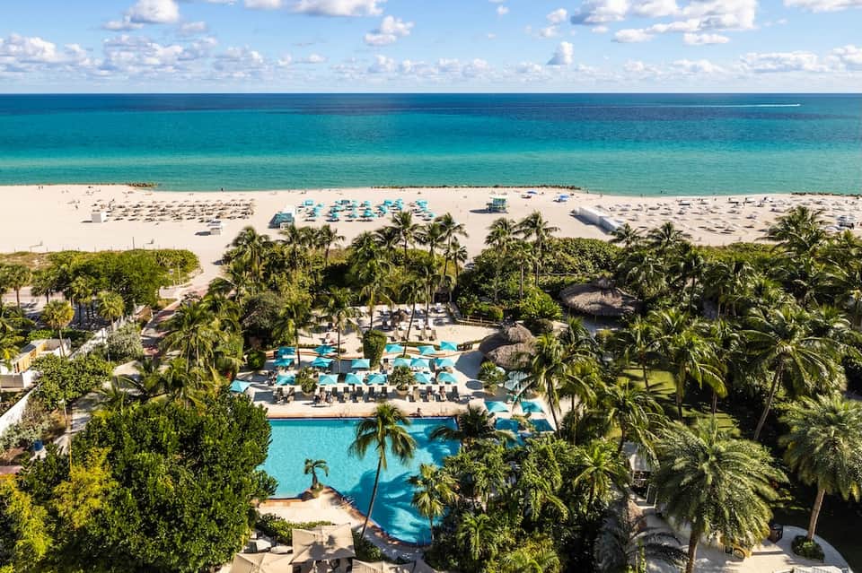 Aerial view of tropical beachfront resort with pools, palm trees, white sand beach, and turquoise ocean