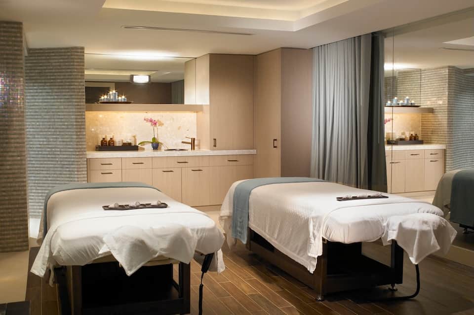Spa treatment room with twin massage tables, wooden cabinetry, mirrors, and ambient lighting