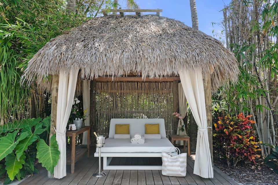 Luxury outdoor cabana with thatched roof, white bed, curtains, and tropical garden surroundings