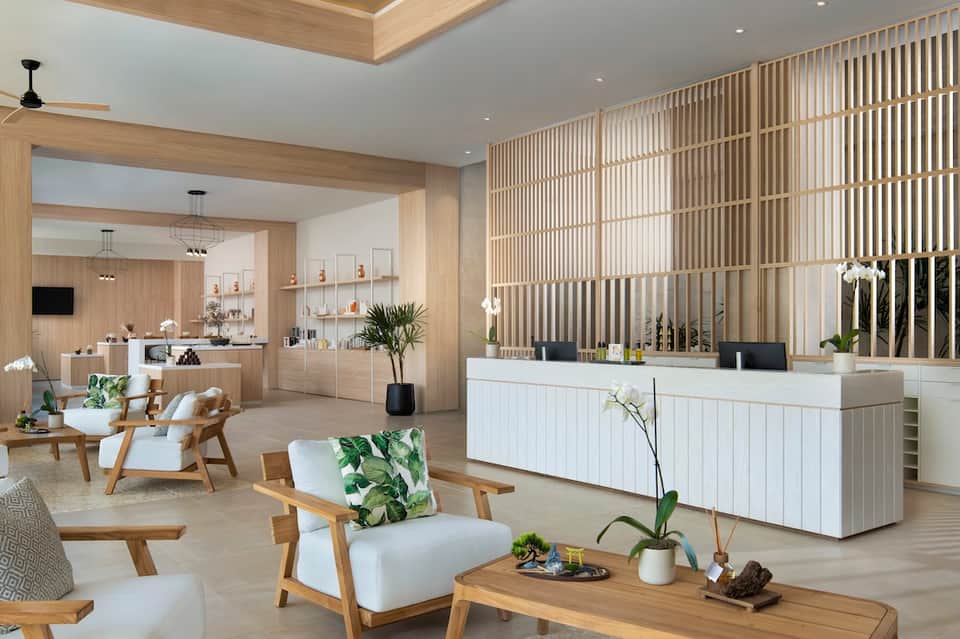 Spa reception area with light wood finishes, white reception desk, vertical slat screens, and plants