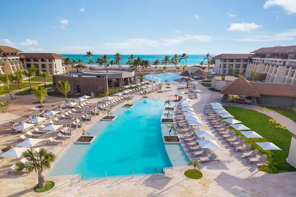Large resort pool complex with multiple levels, lounge chairs, ocean view, and buildings