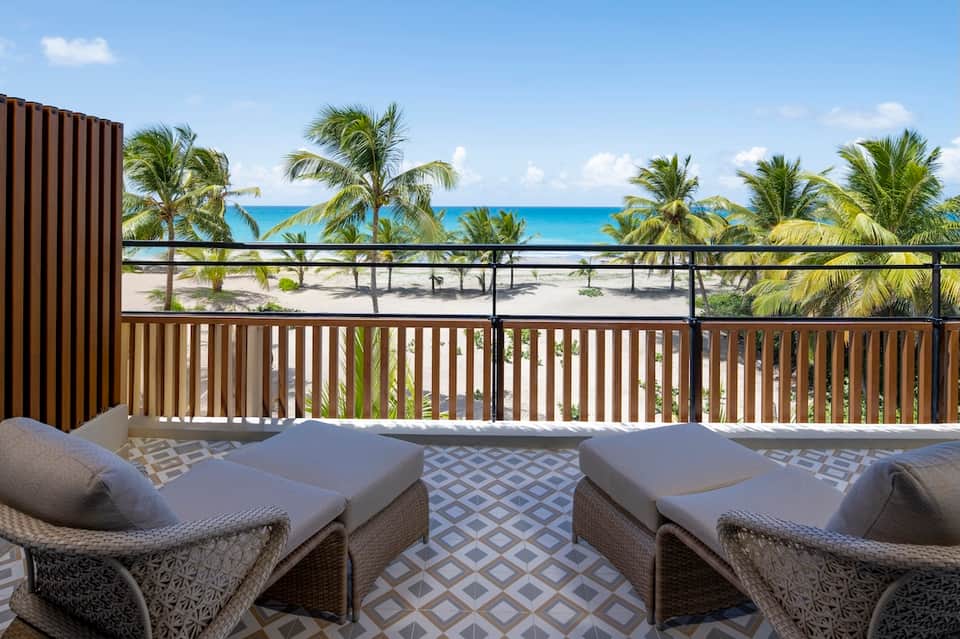 Beachfront balcony with lounge chairs overlooking palm trees, white sandy beach, and clear blue ocean water