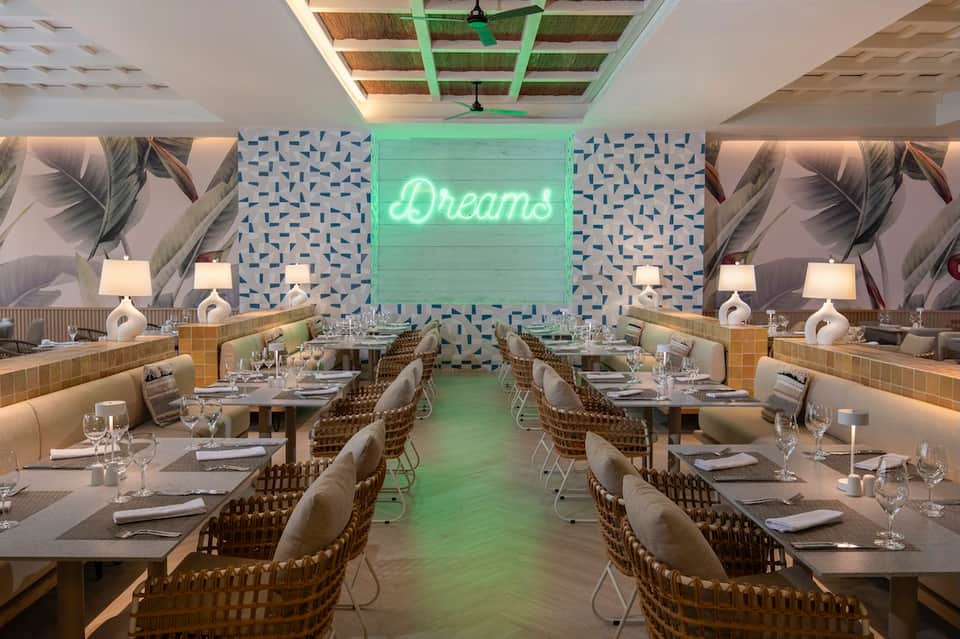 Contemporary dining room with green neon 'Dreams' sign, feather wallpaper, and wooden woven chairs