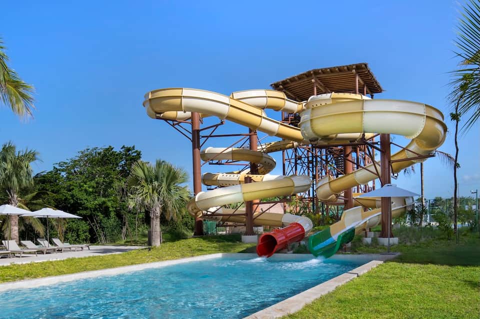 Resort water slide structure with tan tubes, pool below, palm trees, and green lawn