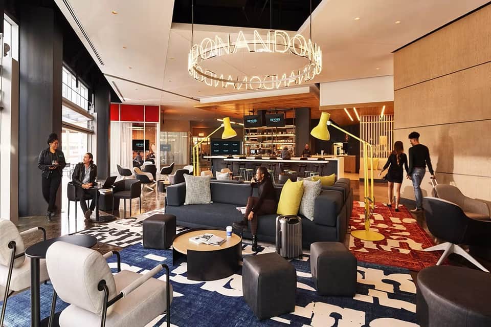 Modern hotel lobby with neon signage, black and gold bar, dark furniture, decorative rugs, and contemporary design
