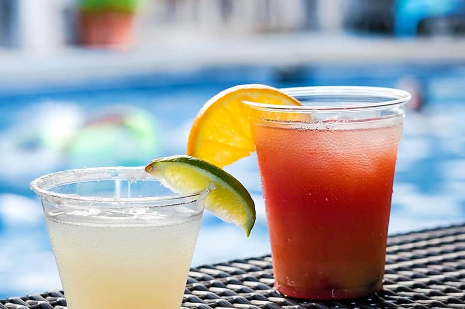 Two colorful cocktails by a pool—a clear drink with lime and a red drink with orange slice.