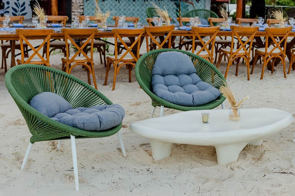 Beach lounge area with green woven pod chairs, white round table, and long dining setup beyond