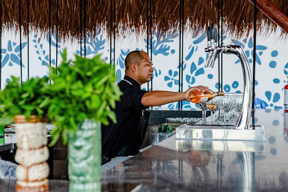 Beachbar bartender pouring rosé wine under thatched roof with blue botanical backdrop