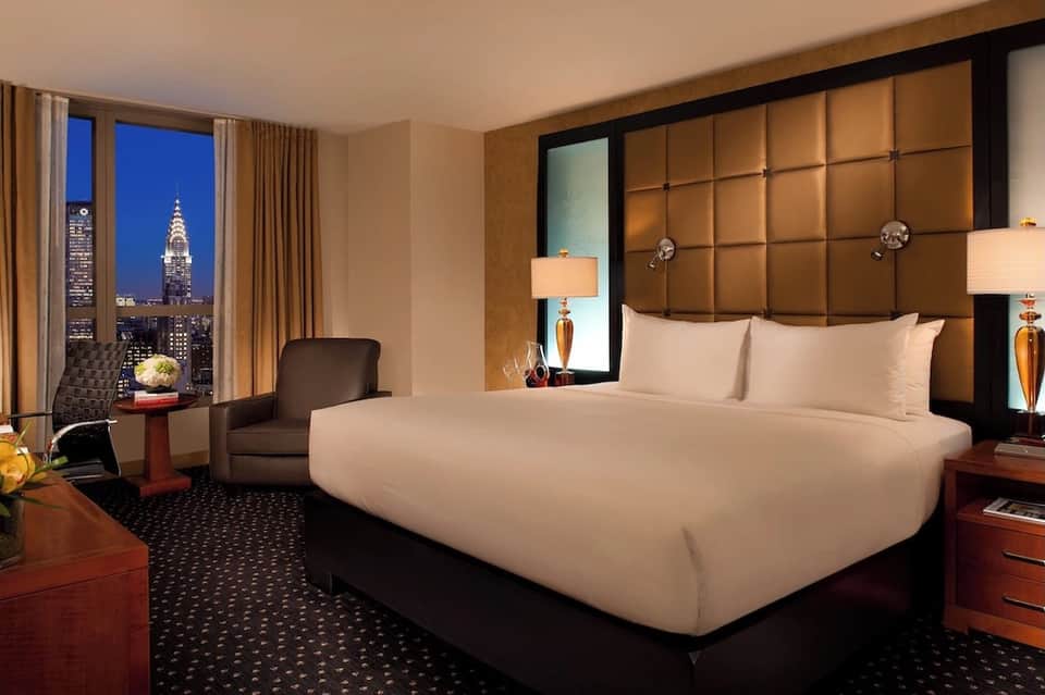Luxury hotel bedroom with king bed, brown leather headboard, city skyline view, and warm lighting