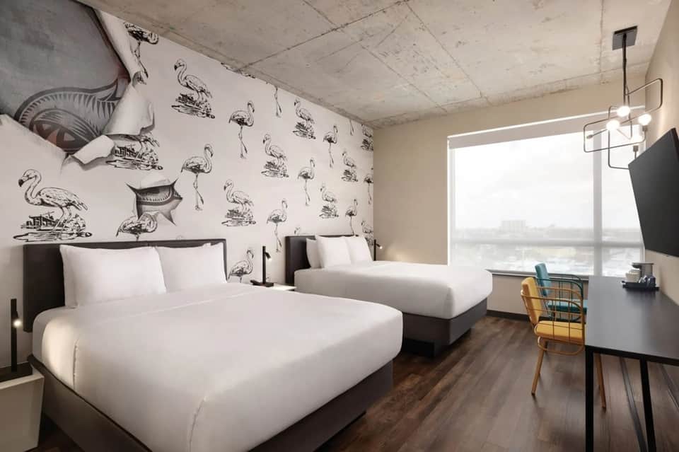 Contemporary hotel room with two beds, flamingo wallpaper, and large window overlooking water