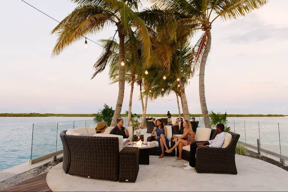 Beachfront fire pit lounge with wicker seating and palm trees overlooking water at dusk