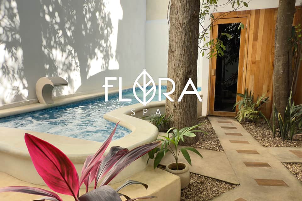 Flora Spa entrance with logo displayed on white wall, hot tub, wooden door, and tropical plants