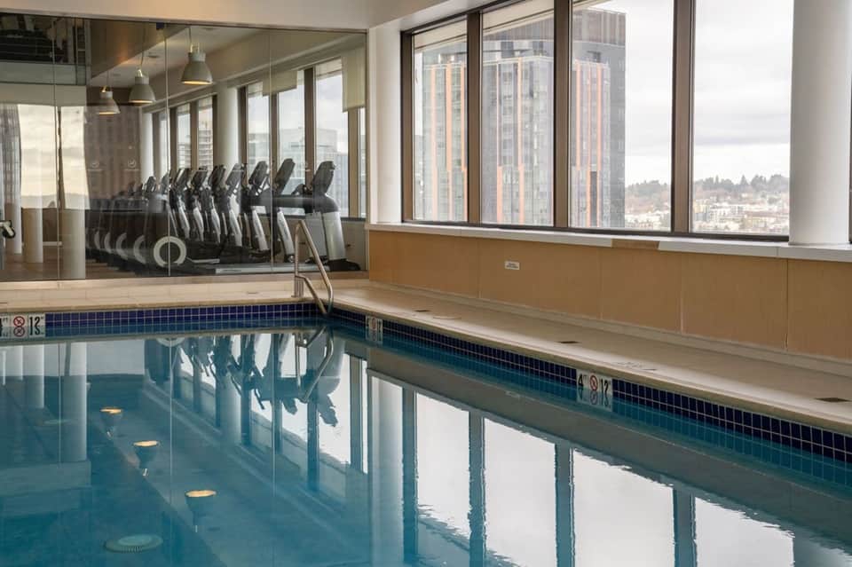 Indoor swimming pool with exercise bikes visible, large windows, and high-rise city skyline views