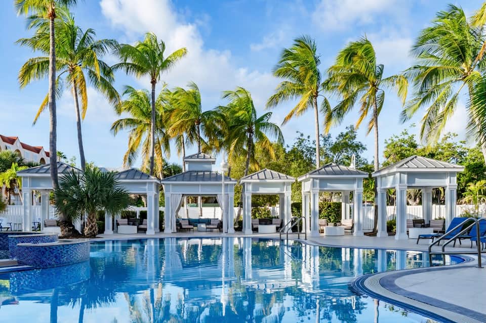 Resort pool with white cabanas, palm trees, and bright blue water reflecting sky