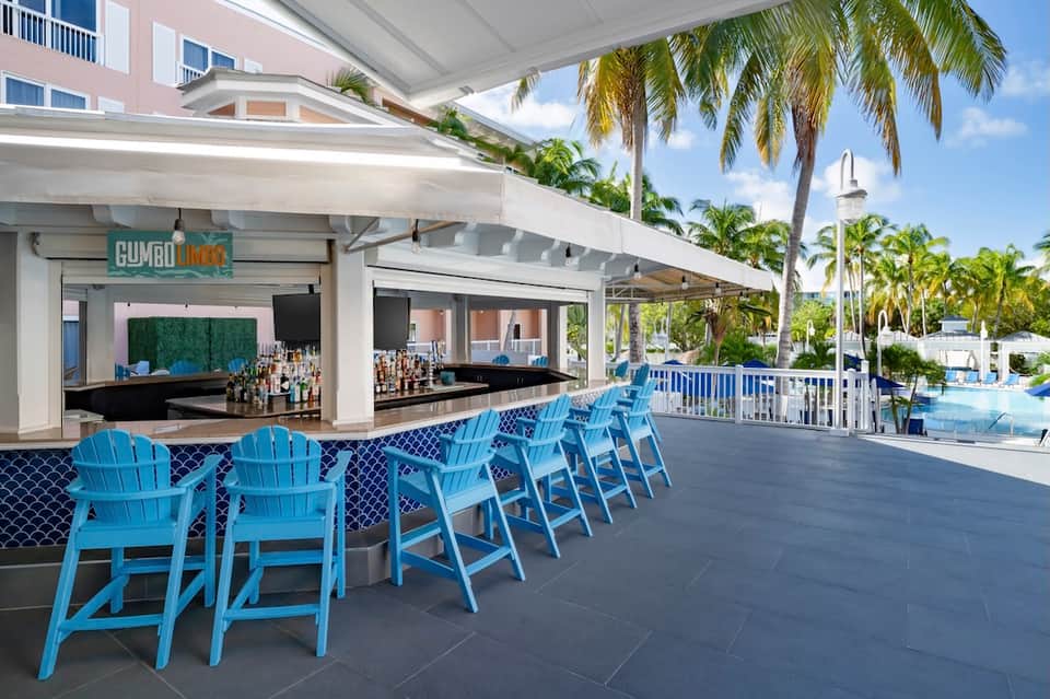 Poolside bar with blue Adirondack chairs, palm trees, and covered terrace seating area